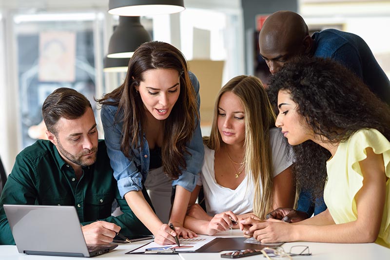 Five young people studying on white desk. Beautiful women and men working together wearing casual clothes. Multi-ethnic group.